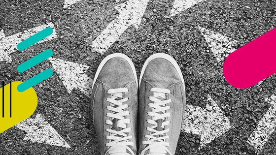 Öffnet die Seite "Schwerpunkt Digitale Beteiligungsprozesse". Beschreibung: Bild in schwarz und weiß mit eingefügten bunten Gestaltungselementen. Man sieht ein Paar Schuhe von oben die auf Asphalt stehen. Auf dem Asphalt sind weiße Pfeile, die in unterschiedliche Richtung zeigen.