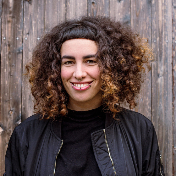 Porträt Nushin Yazdani. Ausschnitt von Kopf bis Brust. Im Hintergrund ist eine Holzwand. Sie trägt eine Lederjacke, lacht und schaut in die Kamera