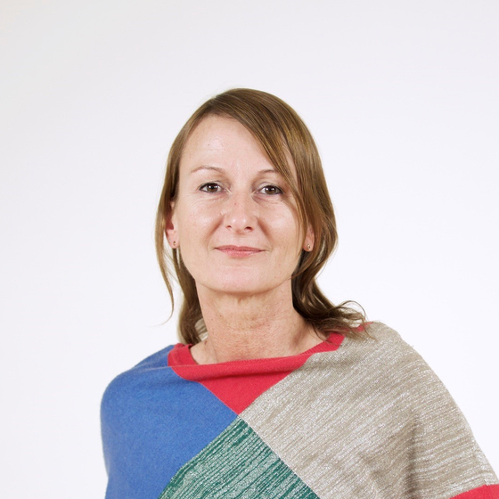 Porträt Sonja Reichmann. Ausschnitt von Kopf bis Hüfte mit einfarbigen Hintergrund. Sie hat die Hände an der Hüfte, trägt einen bunt karierten Pullover und schaut lächelnd in die Kamera.