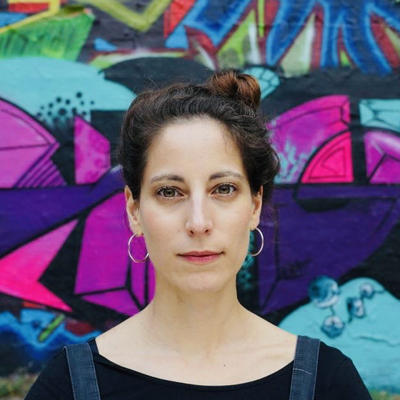 Porträt von Nil Idil Çakmak von Kopf bis Schultern mit Graffiti im Hintergrund. Sie blickt frontal in die Kamera. 